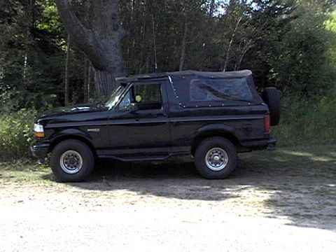 1995 Ford Bronco - A black Ford Bronco with a soft top is parked outdoors on a dirt surface.
