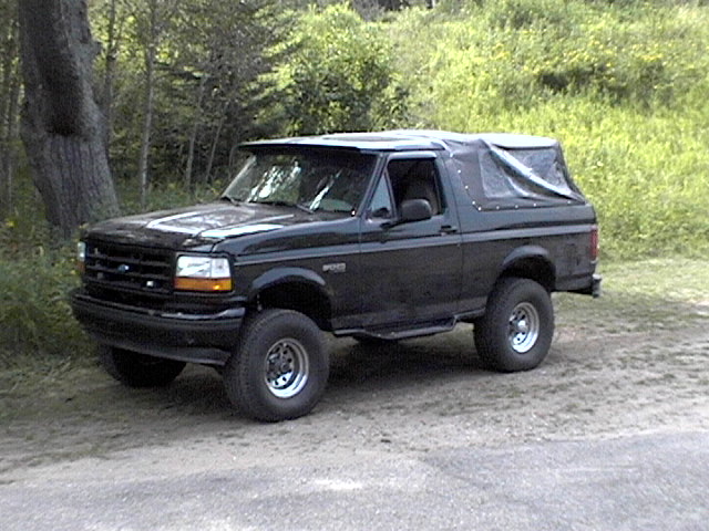 Black Ford Bronco with a soft top parked on a dirt road next to trees