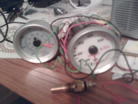 1997 Ford Probe - Two aftermarket white-face gauges with chrome bezels sitting loose on a wood table, their green, pink and black wiring pigtails still uncoi…
