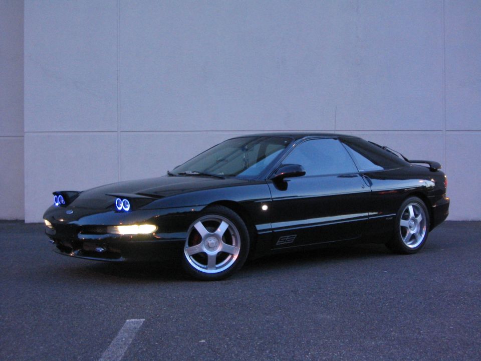 Black 1994 Ford Probe parked in front of a building at dusk. It has pop-up headlights and a spoiler