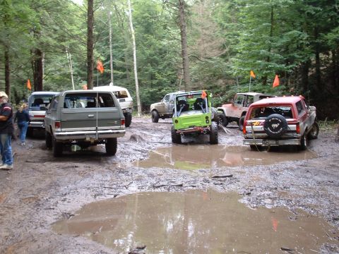 1995 Ford Bronco - Multiple off-road vehicles are gathered in a muddy clearing within a wooded area.
