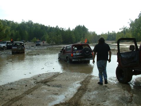 1995 Ford Bronco - Multiple off-road vehicles navigate a muddy terrain with trees in the background.
