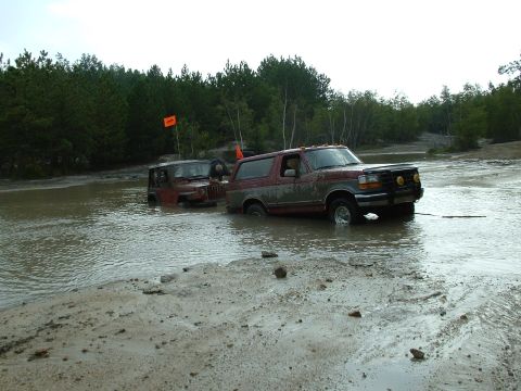 1995 Ford Bronco - A mud-coated red full-size Ford Bronco sits axle-deep in a flooded gravel pit alongside a red Jeep Wrangler that is buried deeper in the wa…