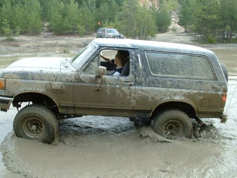 1995 Ford Bronco - A mud-caked full-size Ford Bronco with its upper doors removed churning through a deep muddy water hole, side profile, with the driver visi…