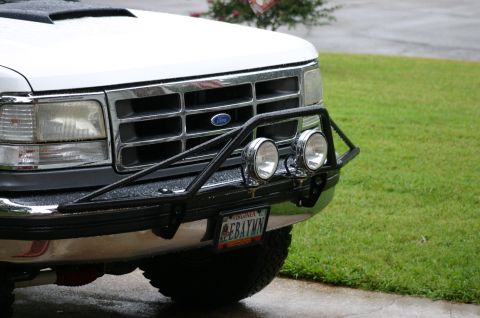 1994 Ford Bronco - Close-up 3/4 front view of a white Ford truck/SUV with a chrome grille bearing the blue Ford oval, fitted with a black tubular brush guard/…