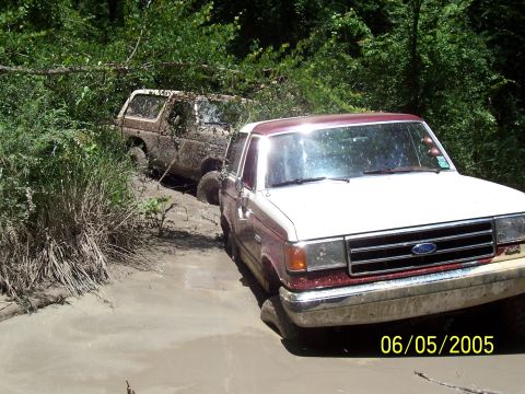 1987 Ford Bronco - Two 1987 Ford Broncos are stuck in a muddy off-road trail, with one vehicle partially submerged.