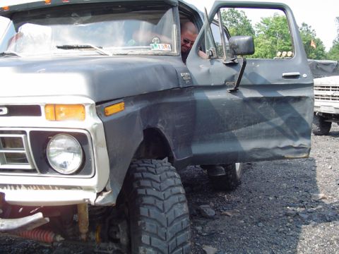 1979 Ford Bronco - A dark-colored pickup truck with its door open, showing a driver inside. Another white truck is visible in the background.