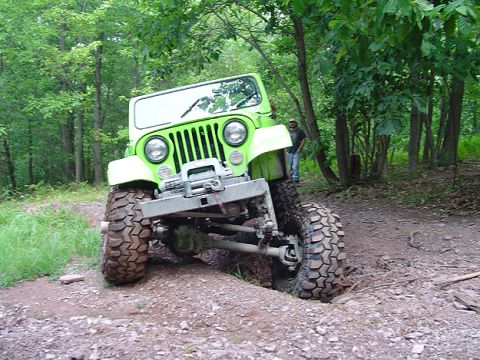 1979 Ford Bronco - A modified green Jeep navigates a challenging dirt trail surrounded by trees.