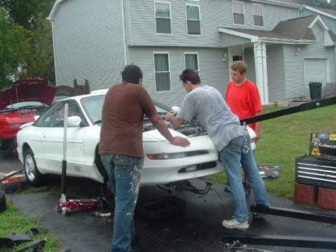 1996 Ford Probe - Three men work on the engine of a white 1996 Ford Probe, with a red car visible in the background.
