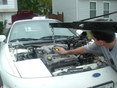 1996 Ford Probe - A person works on the engine of a white 1996 Ford Probe, which is being lifted by an engine hoist.