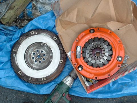 1996 Ford Probe - A new orange clutch assembly and an old metal flywheel are laid out on a blue tarp next to a drill.
