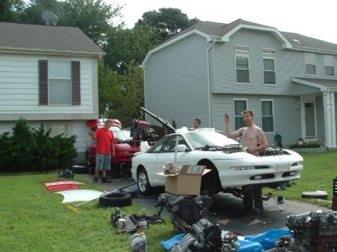 1996 Ford Probe - Two men appear to be in the middle of an engine swap on a white car, with a red car and engine parts scattered around.