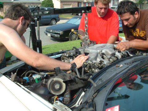 1996 Ford Probe - Three men work on an engine, with a black car and a blue SUV visible in the background.