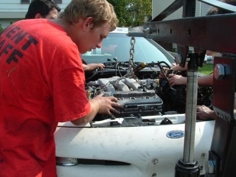 1996 Ford Probe - Mechanics work to remove the engine from a white vehicle, with a Ford emblem visible on the hood.