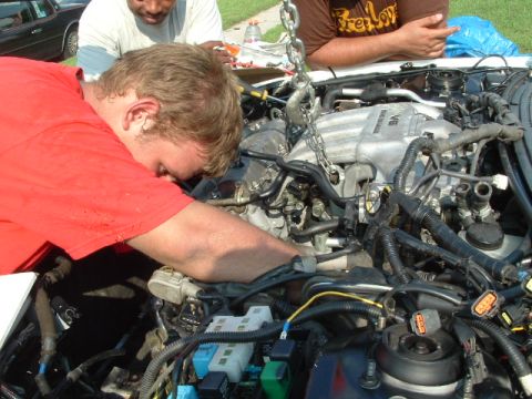 1996 Ford Probe - Mechanics work on a V6 engine in an engine bay, with a chain hoist suspended above.