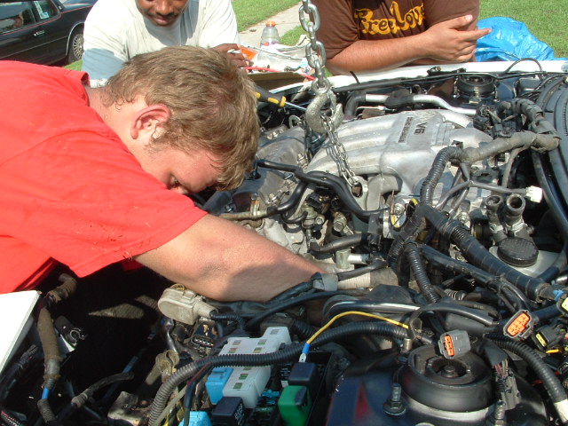 Engine bay with V6 engine being worked on by people, chain hoist attached