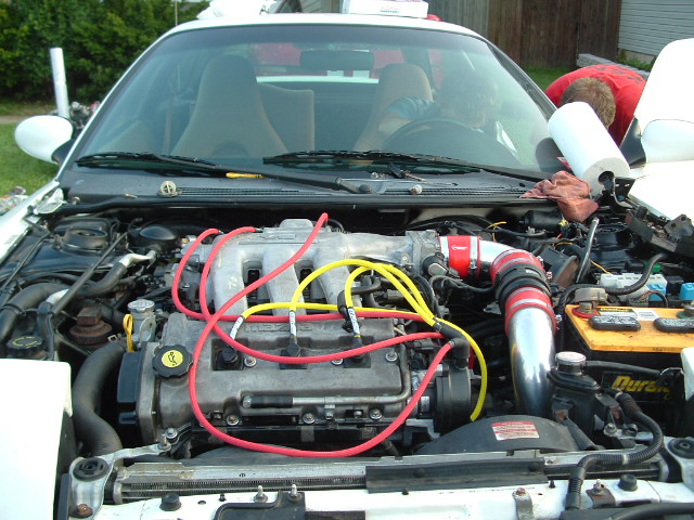 Engine bay of a white car with red and yellow spark plug wires, custom intake piping, and a Duracell battery