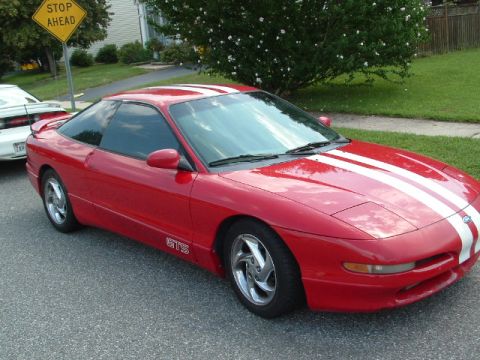 1996 Ford Probe - A red 1996 Ford Probe with white racing stripes and a 'GTS' badge is parked on a street next to another white vehicle.