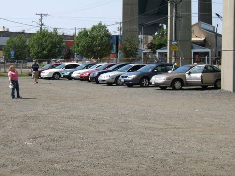 1998 Ford Taurus - Several sedans are parked in a gravel lot under a large bridge structure.