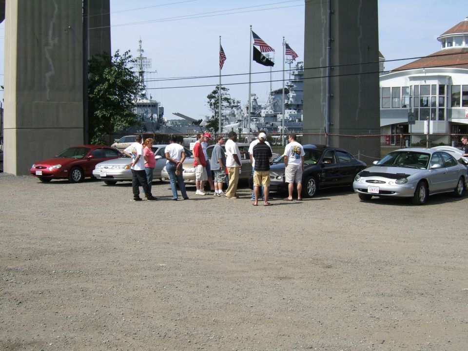Cars parked near a waterfront with ships in the background. People are standing around