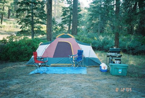 1990 Ford Bronco - large multi-room dome tent stands with red and blue camp chairs, a cooler, and a stove in a sunny pine campsite, dated August 12, 2005.