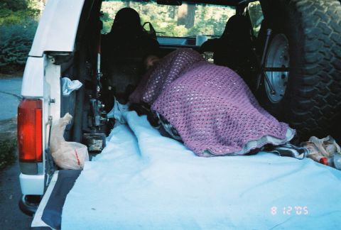 1990 Ford Bronco - person sleeps under a purple crocheted blanket in the open rear cargo area of an SUV, a spare tire visible, dated August 12, 2005.