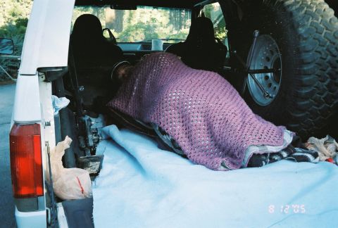 1990 Ford Bronco - person rests under a purple crocheted blanket in the open tailgate cargo bay of an SUV beside a mounted spare tire, dated August 12, 2005.