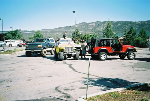 1990 Ford Bronco - lifted full-size SUV, an open CJ-style Jeep and a red Jeep Wrangler sit on a paved lot beneath pine-covered mountains.