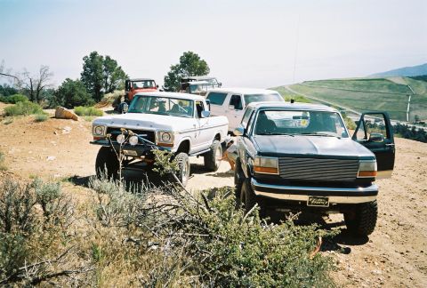 1990 Ford Bronco - lifted pickup with a billet grille and another lifted SUV sit on a dirt ridge with green hills behind.