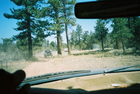 1990 Ford Bronco - Seen through the windshield, a vehicle ahead raises dust on a dirt trail winding through tall pine trees.
