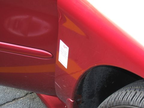 1998 Ford Taurus - Close-up of a red car's side panel and fender, revealing a dent and a sticker with text.