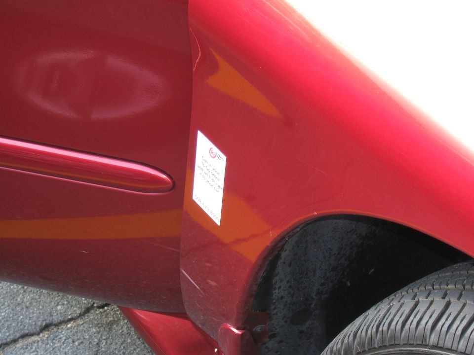 Red car door and fender with a white sticker, showing a dent and scratches