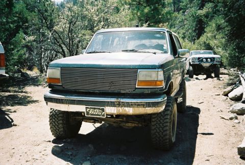 1990 Ford Bronco - muddy lifted Ford full-size SUV with a billet grille faces the camera on a dirt trail with another truck behind.