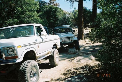 1990 Ford Bronco - white lifted SUV and a gray Jeep Wrangler pass over rocks on a shaded forest trail, dated August 2005.