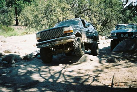 1990 Ford Bronco - lifted dark Ford Bronco flexes its suspension over rocks on a sandy forest trail with a second truck behind.