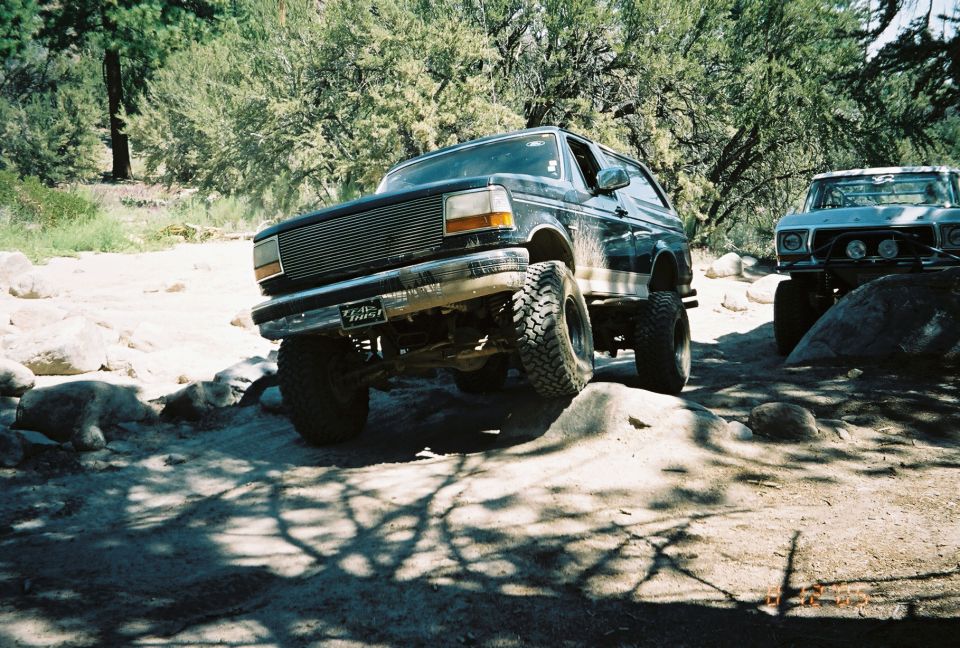Lifted dark Ford Bronco flexing over rocks on a sandy trail with another truck behind
