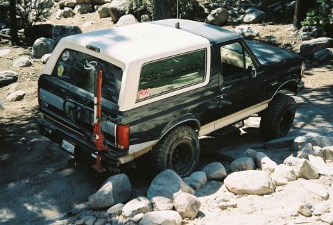 1990 Ford Bronco - black lifted Ford Bronco with a white hardtop and rear-mounted high-lift jack sits among large boulders.
