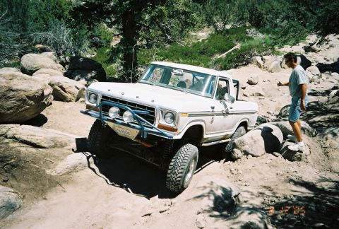 1990 Ford Bronco - white lifted Ford pickup with FORD hood lettering crawls over rocks as a man in shorts watches, dated 2005.