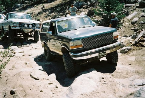 1990 Ford Bronco - lifted dark Ford Bronco and a white vintage pickup sit on a sandy rocky trail as several people look on.