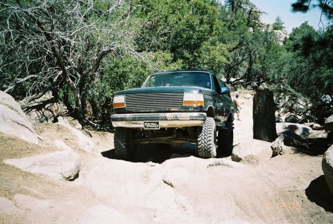 1990 Ford Bronco - dark lifted Ford pickup with a billet grille crawls a sandy rutted trail surrounded by dry brush.