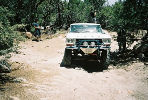 1990 Ford Bronco - white lifted vintage pickup with auxiliary lights and a front license plate crawls a sandy trail as people watch.