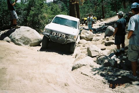 1990 Ford Bronco - white lifted SUV crawls over large white boulders as bystanders watch, with a yellow vehicle on the trail behind.