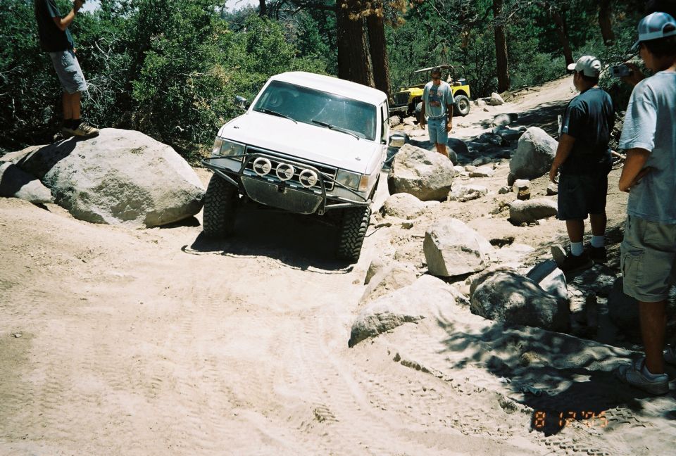 White lifted SUV crawling over large boulders with several bystanders and a yellow vehicle behind