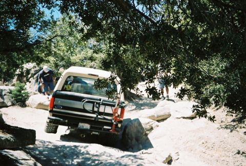 1990 Ford Bronco - lifted Ford full-size SUV with a Ford tailgate emblem crawls over boulders on a tree-shaded off-road trail.