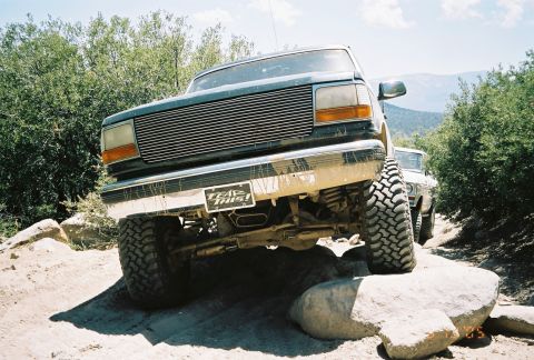 1990 Ford Bronco - muddy lifted Ford truck with a billet grille and a 'Trail This!' license plate flexes over a rock on the trail.