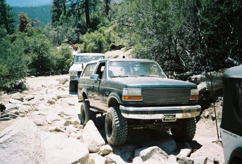 1990 Ford Bronco - lifted dark Ford Bronco on oversized tires sits on a rocky mountain trail among other off-road vehicles.