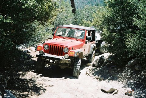 1990 Ford Bronco - red Jeep Wrangler with a folded soft top crawls up a rocky dirt trail through wooded terrain.