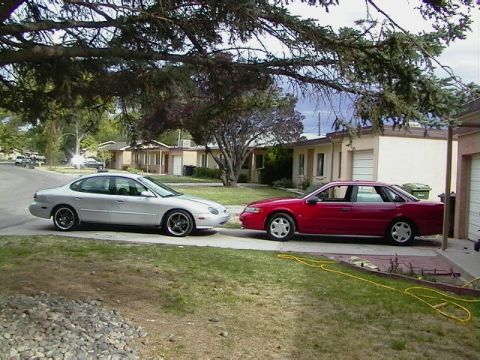 1992 Ford Taurus - Two sedans, a silver Taurus and a red car, are parked in a suburban driveway.
