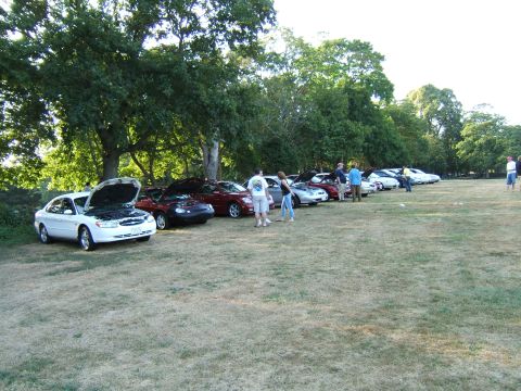 1998 Ford Taurus - A line of cars, including a white 1998 Ford Taurus with its hood up, are parked on a grassy field under trees.