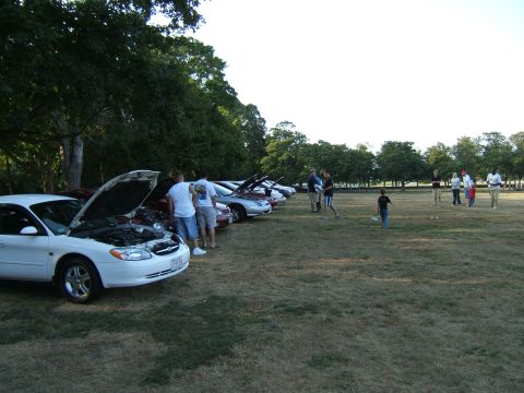1998 Ford Taurus - Several cars are parked on a grassy field with their hoods open, while people gather in the background.
